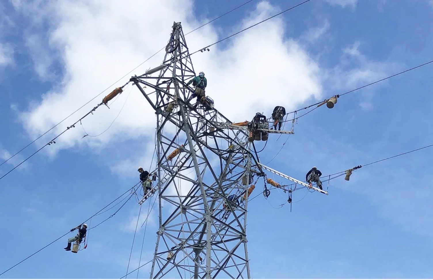 青空の下、鉄塔上で複数の作業員が作業する送電現場